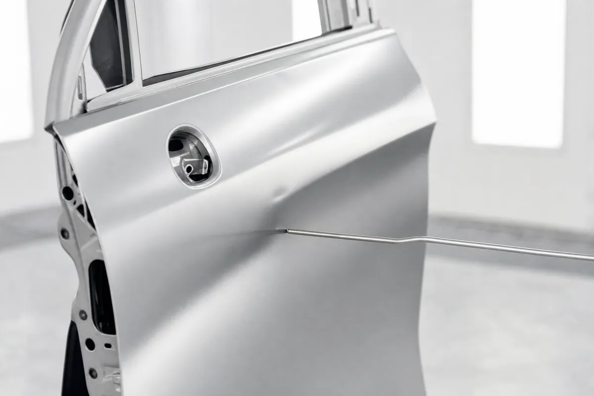 A slim steel dent repair rod reaching in behind a silver door skin, raking light running across the outer panel.